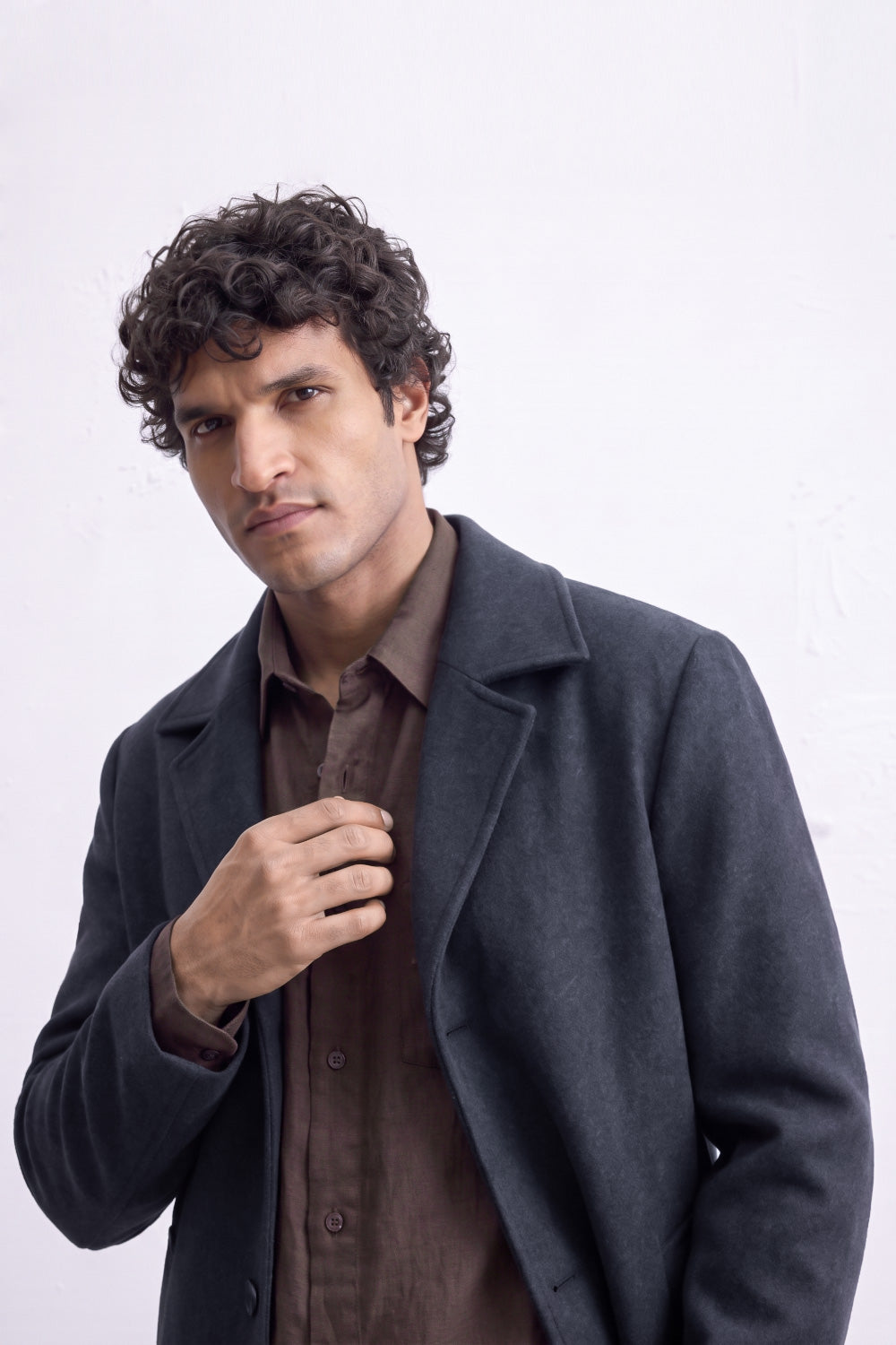 A man with curly dark hair wearing a dark charcoal gray linen coat over a brown button-down shirt, posing against a light off-white background.
