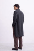 A man wearing a dark charcoal gray mid-length linen overcoat, brown trousers, and black shoes, shown from a side-rear angle against a plain white studio background.