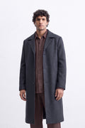A man wearing a long charcoal gray linen overcoat over a brown button-down shirt and brown trousers, standing against a white textured wall.