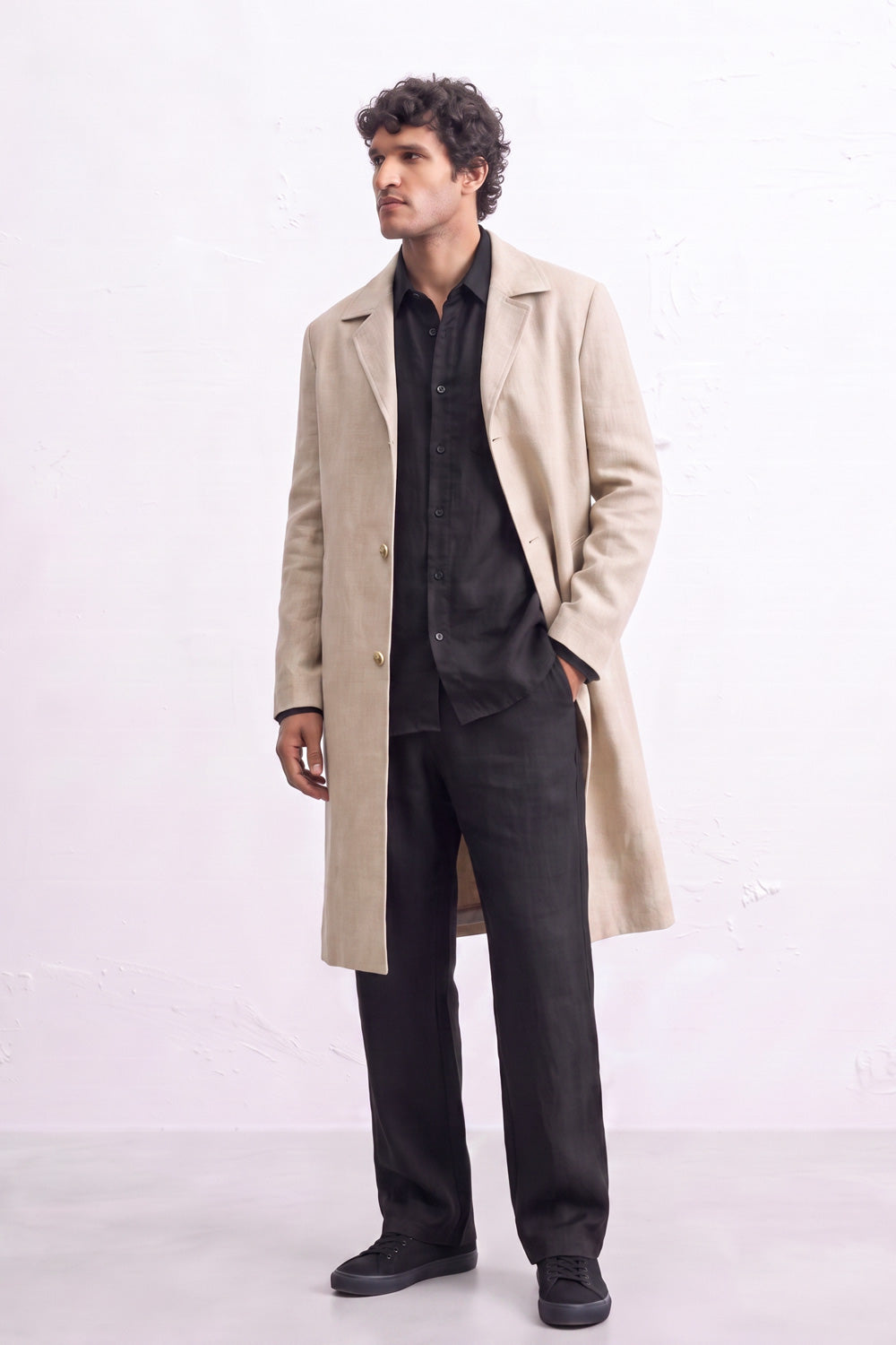 A man modeling a long beige linen trench coat over a black shirt, black trousers, and black shoes, standing against a light off-white textured wall.