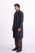 A man wearing a long charcoal gray linen coat over a brown shirt and brown trousers, standing in profile against a white studio background.