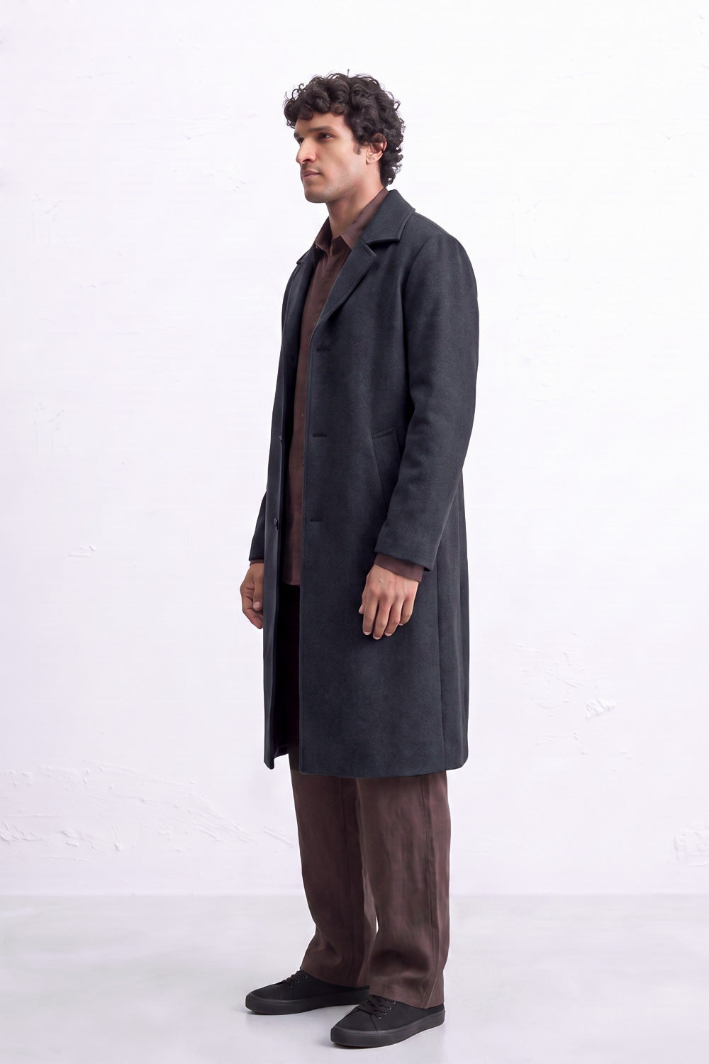 A man wearing a long charcoal gray linen coat over a brown shirt and brown trousers, standing in profile against a white studio background.
