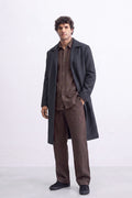 A man modeling a dark charcoal gray linen overcoat over a brown linen shirt and matching brown trousers, standing against a light gray studio background.