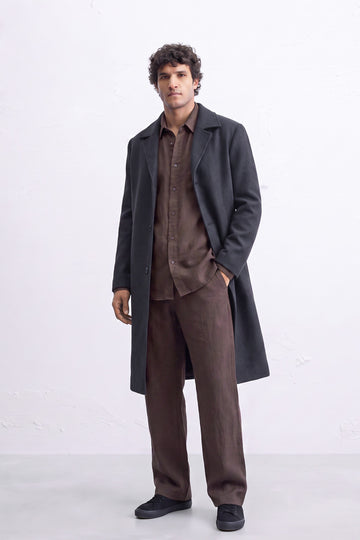 A man modeling a dark charcoal gray linen overcoat over a brown linen shirt and matching brown trousers, standing against a light gray studio background.