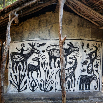 A hand-painted mud wall mural featuring black tribal animal and human figures on a white background, set within a rustic thatched-roof structure.