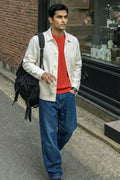 A man walking on a city street wearing a cream-colored linen jacket over a red crewneck shirt, blue jeans, and carrying a black backpack.