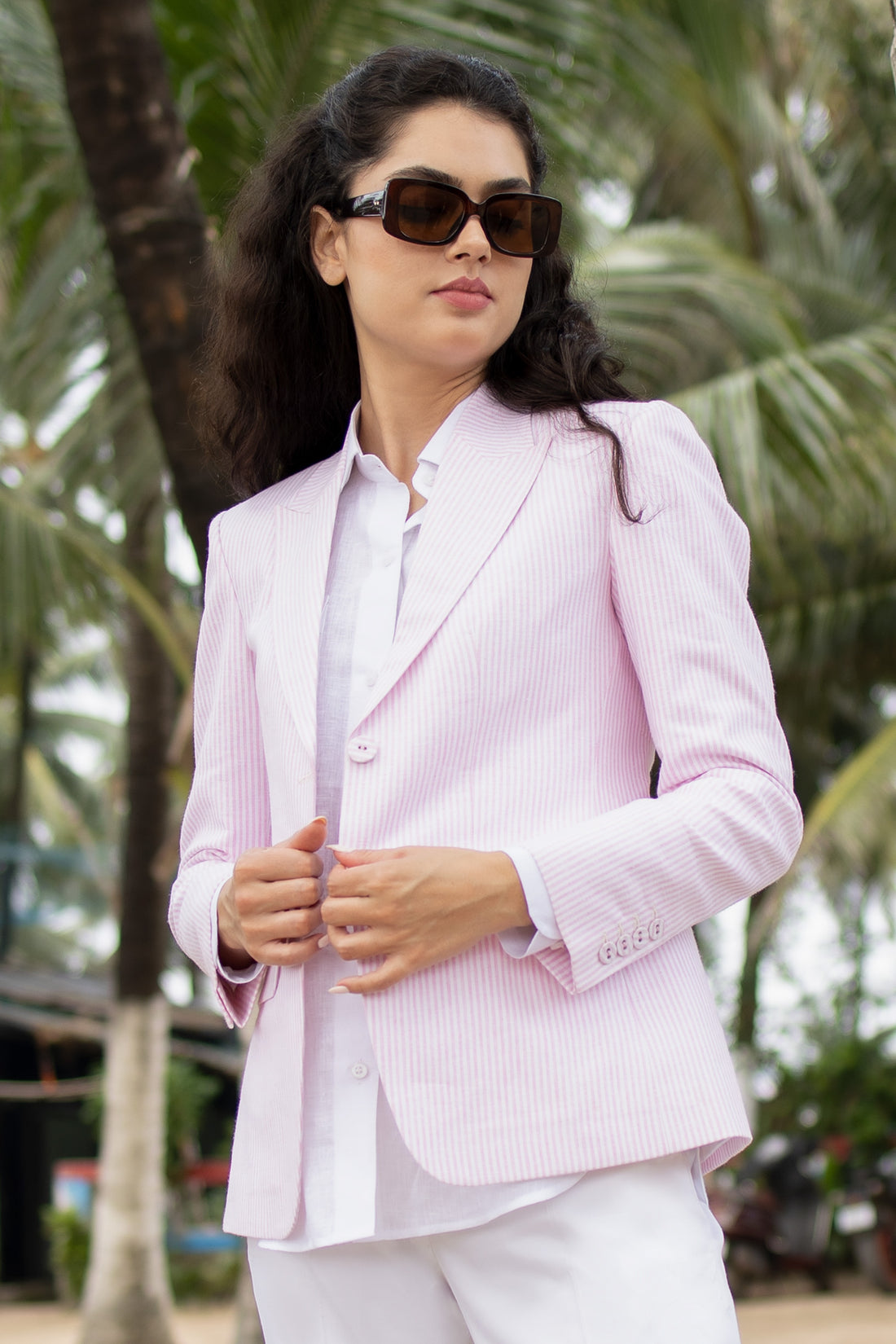 A woman standing outdoors wearing a pink linen blazer with a single-breasted design, peak lapel, and flap pockets. She also has on white trousers and is posing with her hands by her sides.