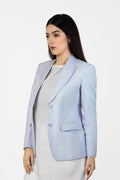 A woman standing outdoors wearing a Sky Blue linen blazer with a single-breasted design, peak lapel, and flap pockets. She also has on white trousers and is posing with her hands by her sides.