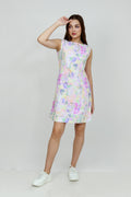 A woman standing in a studio setting, wearing a mid-length floral hemp seersucker dress with a boat neckline and sleeveless top, paired with white sneakers.