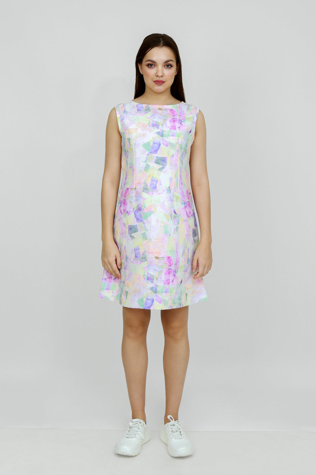 A woman standing in a studio setting, wearing a mid-length floral hemp seersucker dress with a boat neckline and sleeveless top, paired with white sneakers.