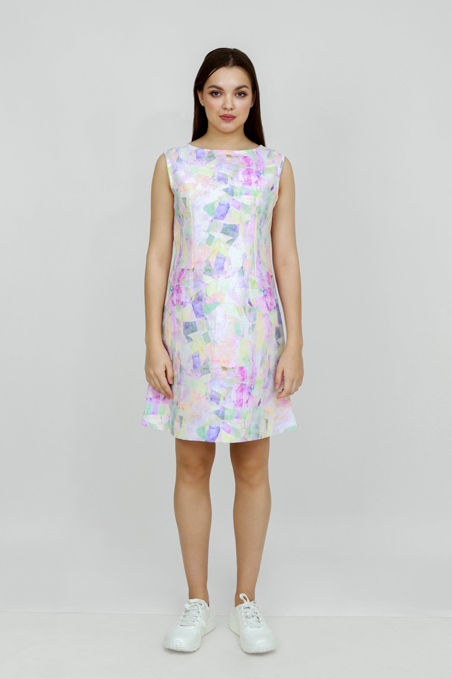 A woman standing in a studio setting, wearing a mid-length floral hemp seersucker dress with a boat neckline and sleeveless top, paired with white sneakers.