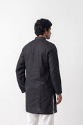 A man viewed from the back wearing a black linen kurta and white trousers, standing against a plain white studio background.