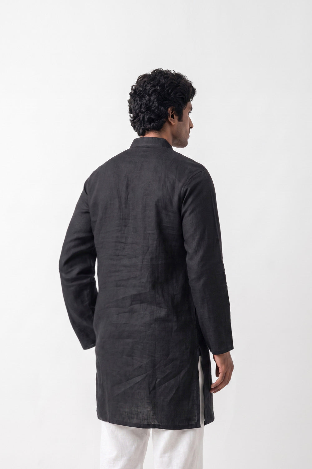 A man viewed from the back wearing a black linen kurta and white trousers, standing against a plain white studio background.