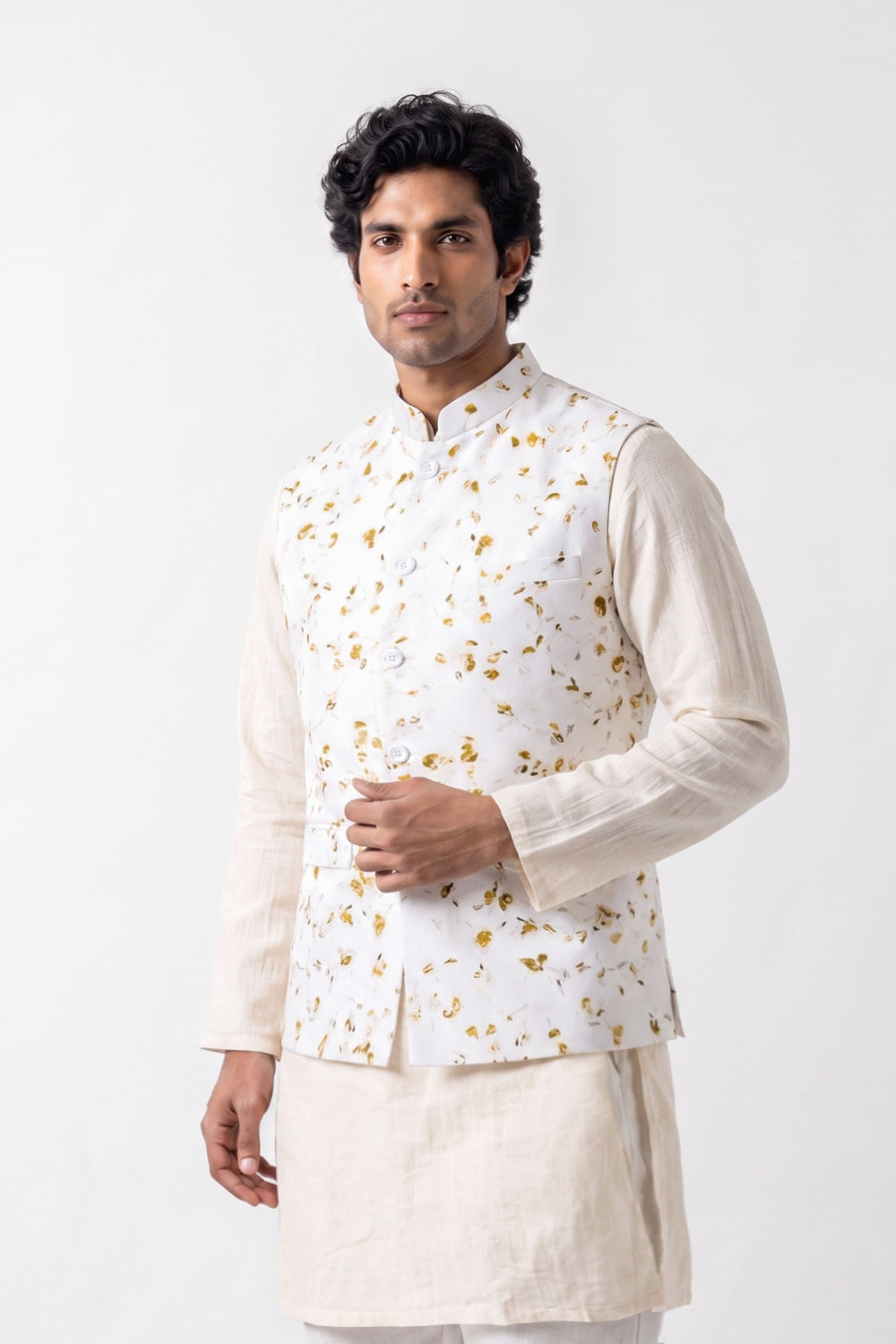 A man wearing a white traditional Indian outfit with a white waistcoat decorated with small gold floral eco-print, paired with a cream linen kurta and trousers, set against a plain white studio background.