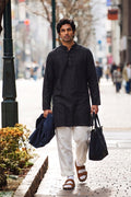 A man walking on a city sidewalk wearing a black long-sleeve linen kurta and white trousers, carrying a dark blue jacket and a black tote bag.