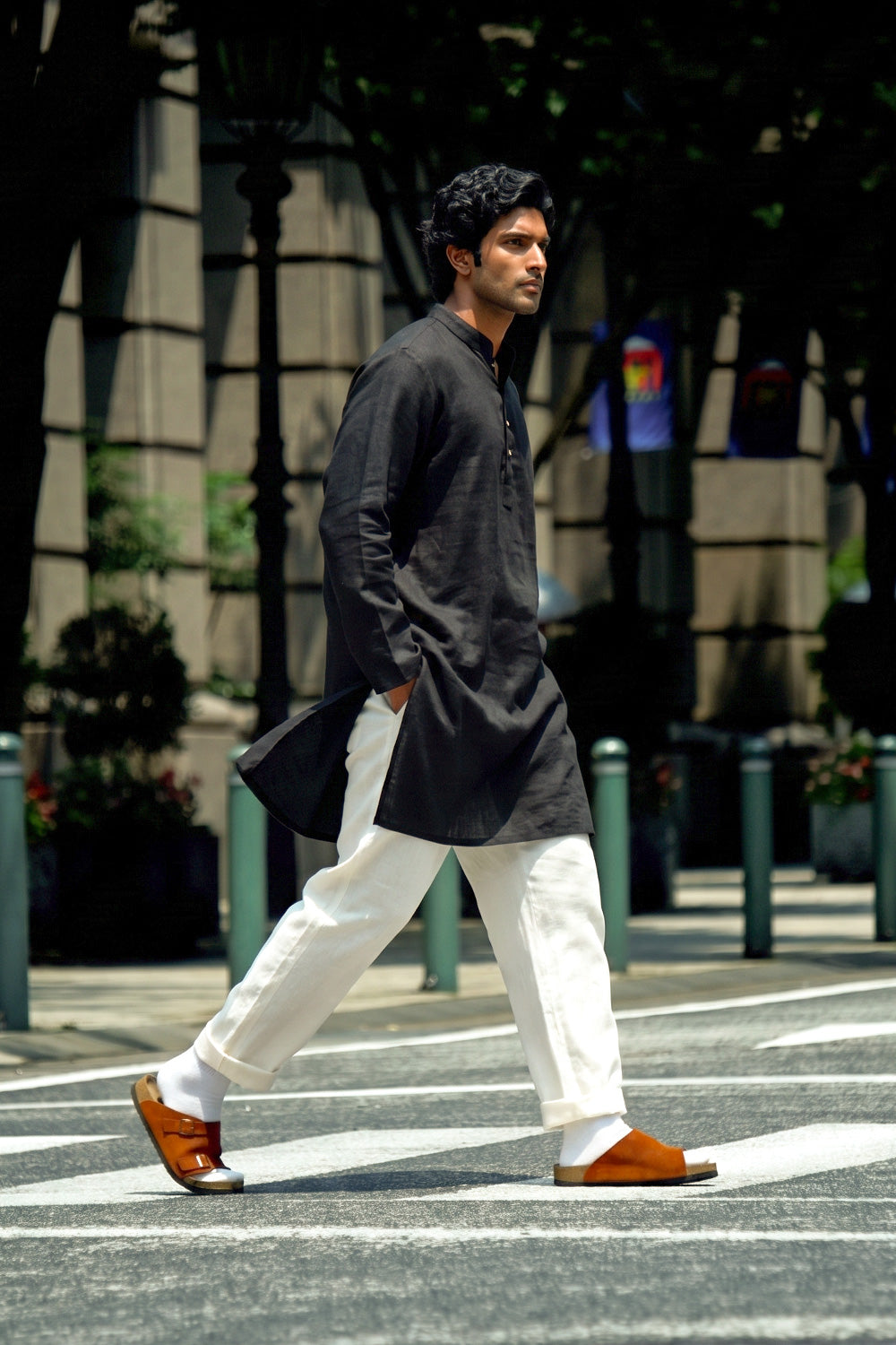A man walking across a city crosswalk wearing a black long-sleeve linenkurta, white trousers, and brown leather sandals.
