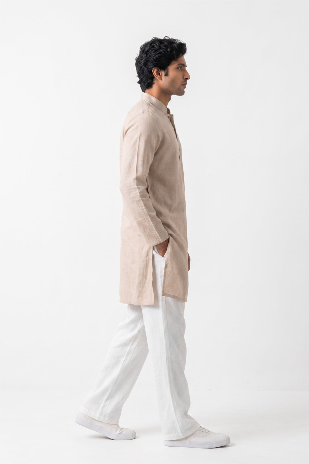 A man in profile wearing a light beige linen kurta and white trousers, standing against a plain white studio background.
