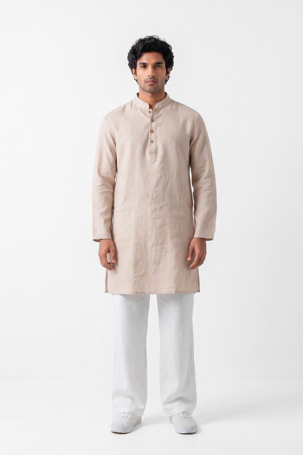 A man modeling a light beige linen kurta with a buttoned mandarin collar, paired with white trousers and white sneakers, standing against a plain white studio background.