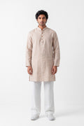 A man modeling a light beige linen kurta with a buttoned mandarin collar, paired with white trousers and white sneakers, standing against a plain white studio background.