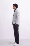 A man in a light gray and white pinstripe linen blazer, black shirt, black trousers, and white sneakers, standing in profile against a white studio background.