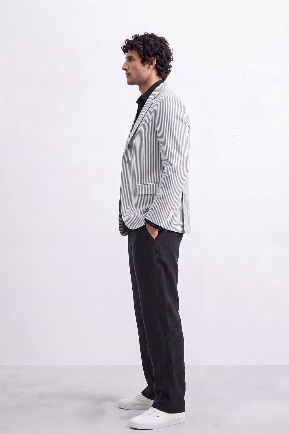 A man in a light gray and white pinstripe linen blazer, black shirt, black trousers, and white sneakers, standing in profile against a white studio background.