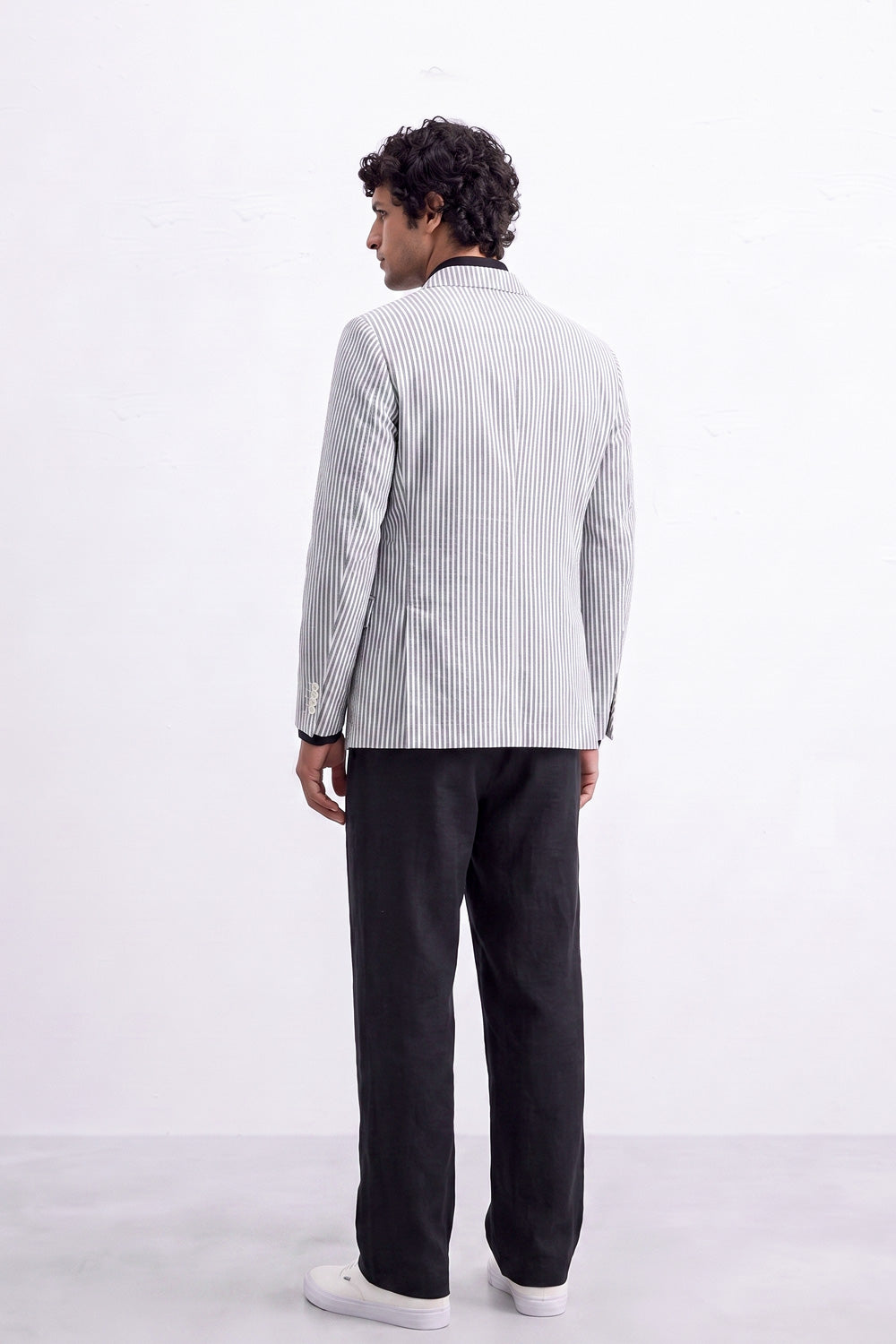 A man shown from the back wearing a light gray and white vertically striped linen blazer, black trousers, and white sneakers, standing against a plain white studio background.