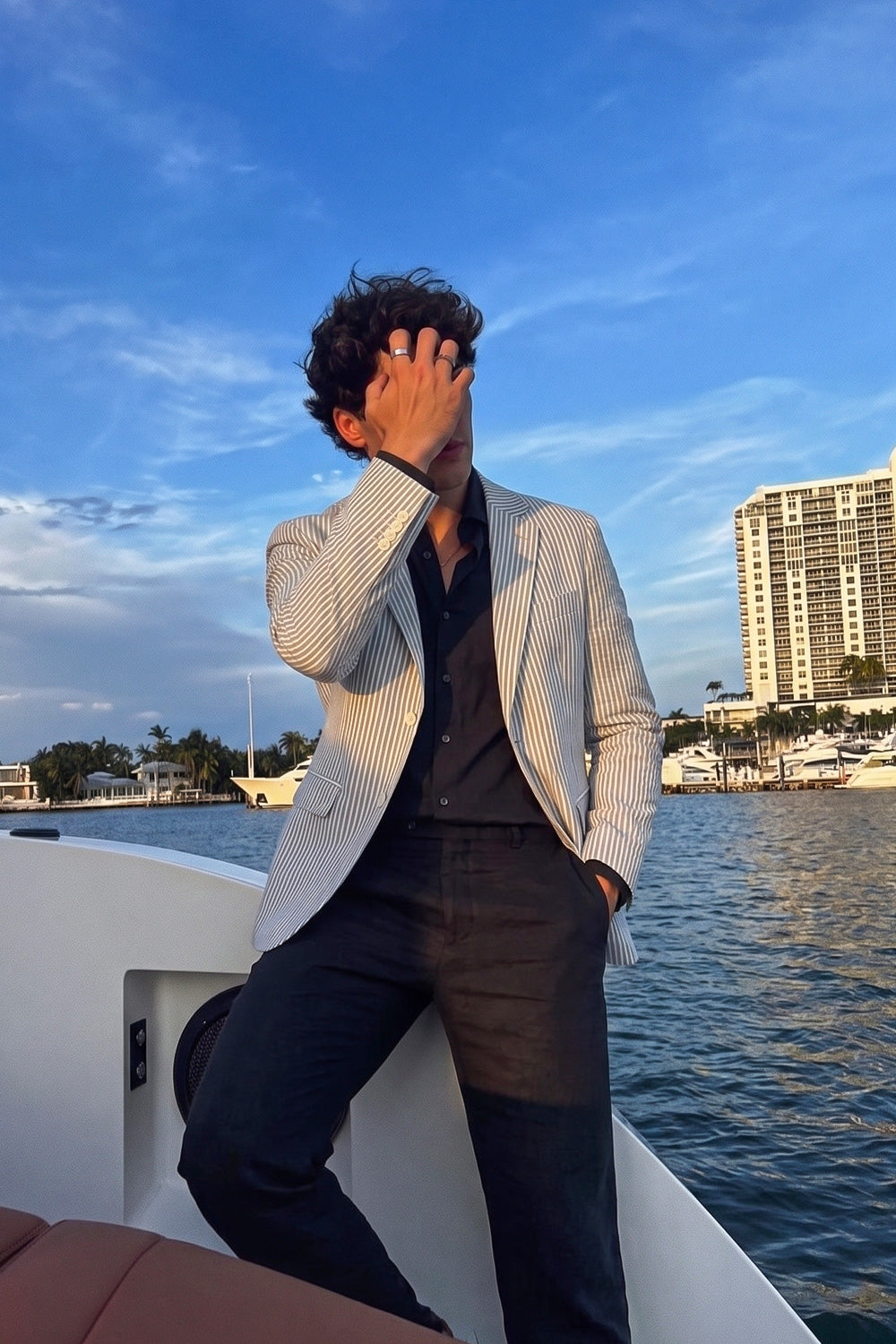 A man posing on a white yacht deck outdoors, wearing a black and white vertically striped linen blazer over a black shirt and dark trousers, with water, a city skyline, and a blue sky in the background.