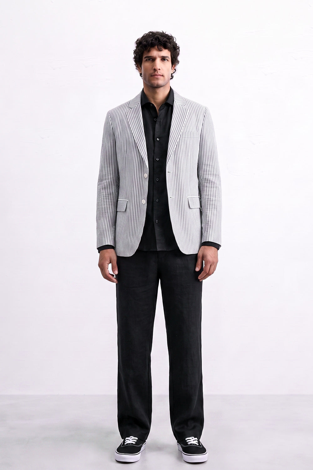 A man modeling a light gray and white vertically striped linen blazer over a black shirt and black trousers, standing against a plain white studio background.