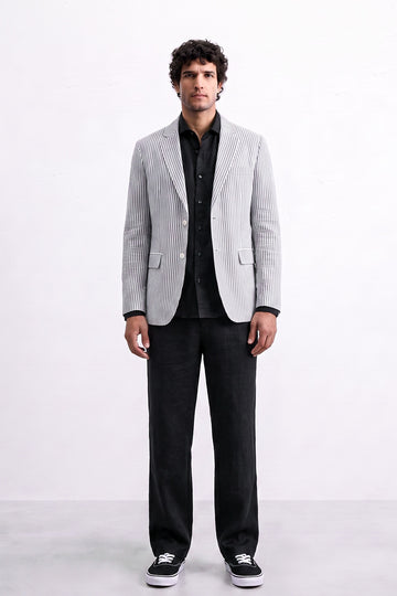 A man modeling a light gray and white vertically striped linen blazer over a black shirt and black trousers, standing against a plain white studio background.