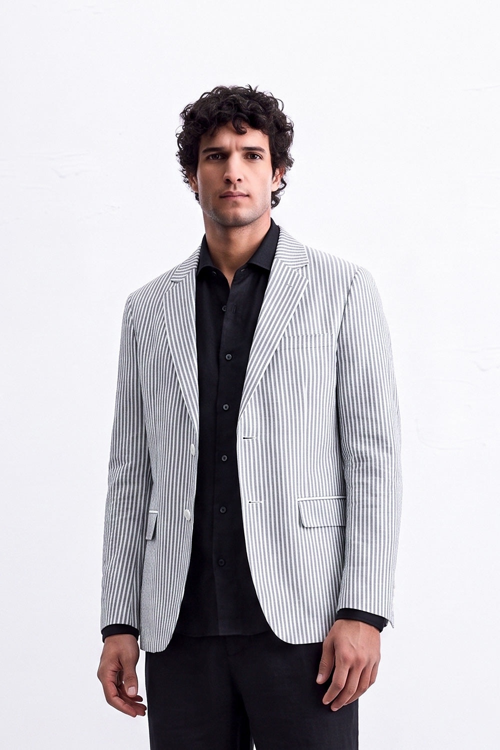 A man modeling a light gray and white vertically striped linen blazer over a black shirt and black trousers, standing against a plain white studio background.