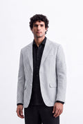 A man wearing a light gray and black pinstriped linen blazer over a black button-down shirt and black trousers, standing against a plain white background.
