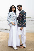 Man wearing a butterfly printed blazer on a wall background a white long shirt, and white trousers. Accessories include sunglasses and a watch.