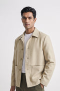 A man wearing a light beige linen overshirt jacket over a white t-shirt and dark olive pants, standing against a light gray studio background.