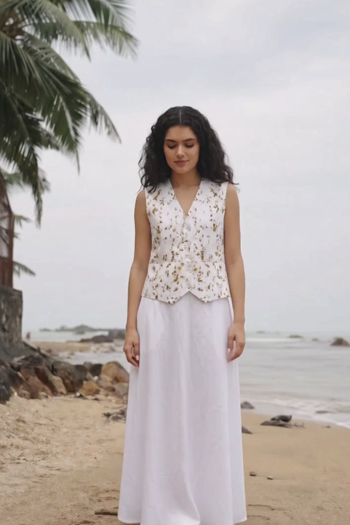 A woman wearing a sleeveless white embroidered vest and a long white skirt with pockets, standing on a sandy beach with water and palm trees in the background.