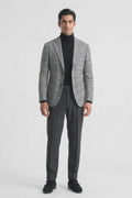 A man modeling a gray and black checks cotton blazer over a black turtleneck, paired with dark gray trousers and black loafers, against a plain white studio background.
