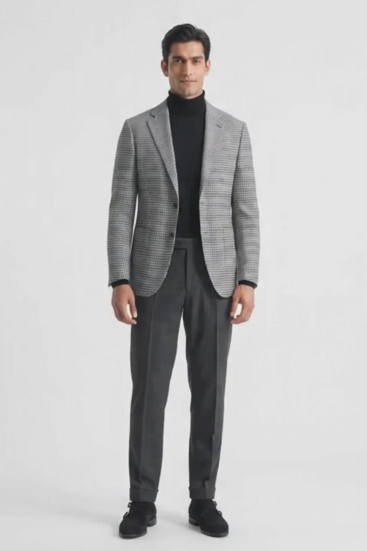 A man modeling a gray and black checks cotton blazer over a black turtleneck, paired with dark gray trousers and black loafers, against a plain white studio background.