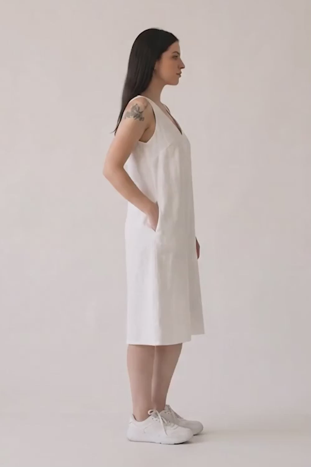 A woman modeling a sleeveless white hemp midi dress with a V-neckline and side pockets, paired with white sneakers, against a plain off-white studio background.