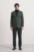 A man wearing a dark olive green linen blazer with flap pockets over a black turtleneck sweater, black trousers, and black leather shoes, standing against a plain white studio background.