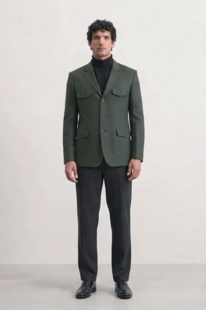 A man wearing a dark olive green linen blazer with flap pockets over a black turtleneck sweater, black trousers, and black leather shoes, standing against a plain white studio background.