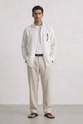 A man modeling a white long-sleeve linen shirt over a white tee, paired with beige linen trousers, a black belt, and black sandals, standing against a light gray studio background.