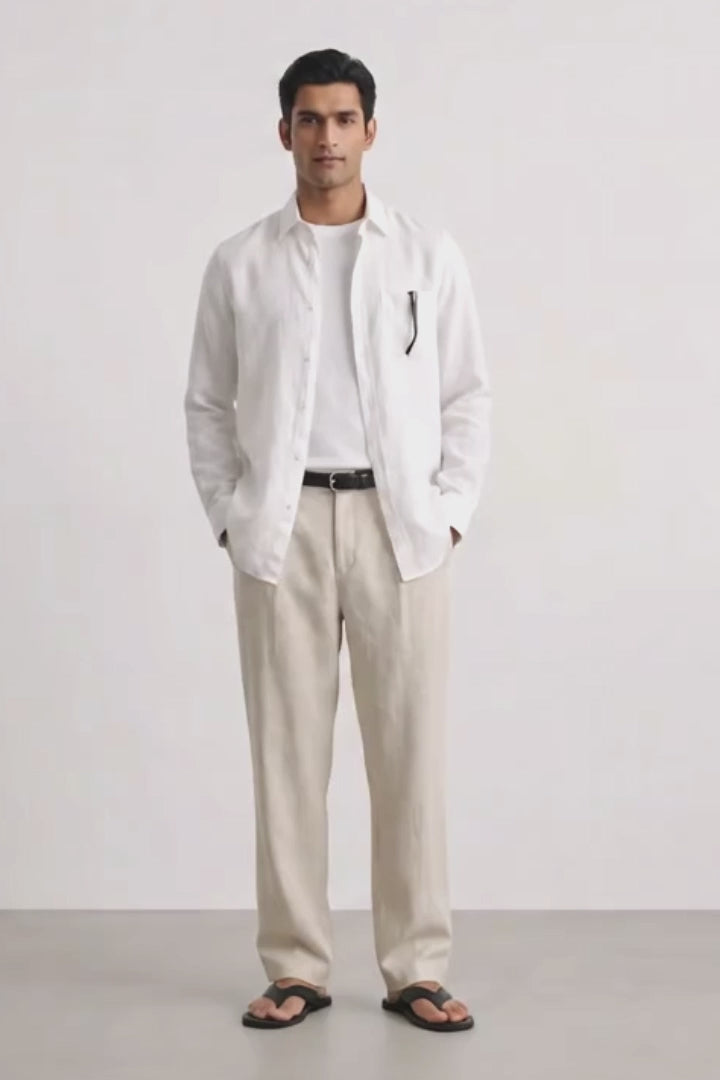 A man modeling a white long-sleeve linen shirt over a white tee, paired with beige linen trousers, a black belt, and black sandals, standing against a light gray studio background.