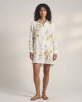 A woman with dark curly hair standing against a plain white background, facing the camera. She is wearing a knee-length, white, long-sleeved button-down shirt dress decorated with yellow and brown botanical prints (leaves and flowers). She is wearing simple flat brown sandals
