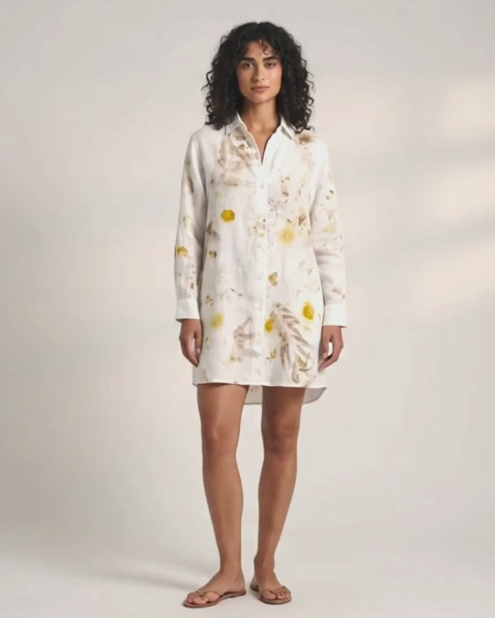 A woman with dark curly hair standing against a plain white background, facing the camera. She is wearing a knee-length, white, long-sleeved button-down shirt dress decorated with yellow and brown botanical prints (leaves and flowers). She is wearing simple flat brown sandals