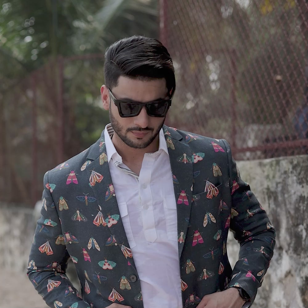 Man wearing a butterfly printed blazer on a wall background a white long shirt, and white trousers. Accessories include sunglasses and a watch.
