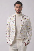 A man modeling a cream-colored blazer with a colorful cocktail glass pattern, paired with a cream turtleneck, cream trousers, and tan suede loafers, against a light gray studio background.
