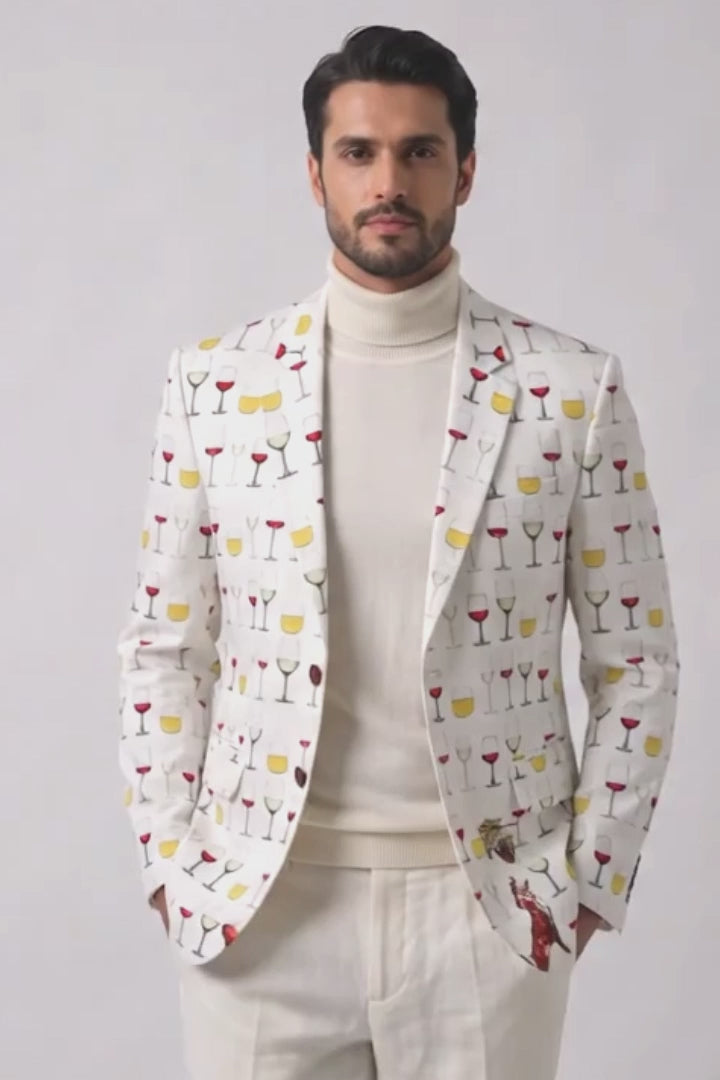 A man modeling a cream-colored blazer with a colorful cocktail glass pattern, paired with a cream turtleneck, cream trousers, and tan suede loafers, against a light gray studio background.