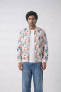 A man modeling a white bomber hemp jacket with a colorful floral print, worn over a white t-shirt and blue jeans, against a plain white studio background.