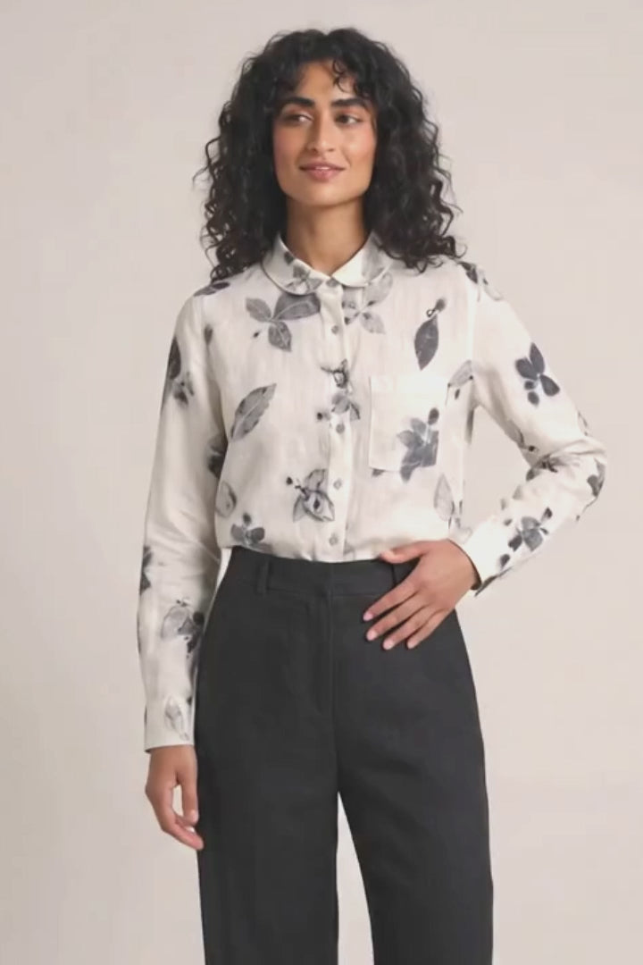 A model stands against a plain light background wearing a white long-sleeve button-up shirt with a gray eco-printed floral pattern, paired with black wide-leg trousers and black shoes.