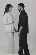 A woman wearing a beige linen vest, shirt, and wide-leg trousers and a man wearing a black vest, long kurta, and trousers as they move around each other against a white background.