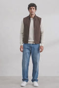 A man modeling a brown sleeveless zippered linen vest over a cream long-sleeve shirt, paired with blue jeans and white sneakers, standing against a white studio wall.